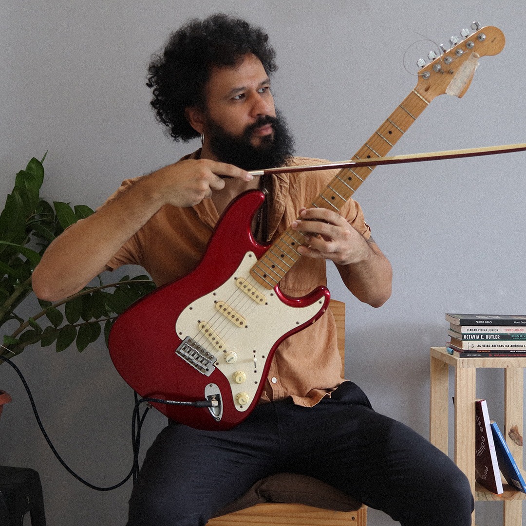 Fotografia colorida. Leandro está sentado em uma cadeira de madeira em uma sala clara, com uma estante de livros a direita e um vaso de plantas a esquerda. Leandro toca as cordas de uma guitarra vermelha e branca que está apoiada na perna esquerda dele. Ele toca a guitarra com um arco que segura na mão esquerda. Leandro tem cabelos blackpower escuros e barba cheia. Usa calça preta e camisa laranja.