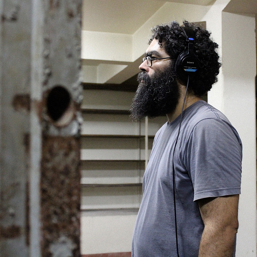 Fotografia colorida. Thelmo está de perfil e usa fones de ouvido supraauriculares pretos. Thelmo está com as duas mãos para baixo e olha para frente. Ele é um homem negro de pele clara, tem o cabelo escuro Black Power, barba grisalha e cheia. Usa uma camiseta cinza e óculos de grau. O fundo da foto é bege e há alguns desníveis na parede.